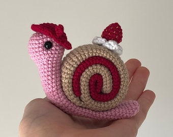 May include: A crocheted snail with a pink body, brown and red shell, and a red hat. The shell is decorated with a white swirl and a red strawberry.