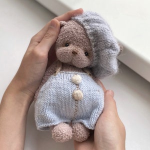 Pode incluir: Um pequeno urso de peluche de malha que usa um chapéu azul de malha e um macacão. O urso tem olhos castanhos e um nariz castanho.