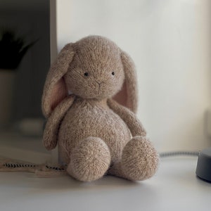 Könnte beinhalten: Ein weiches, braunes, gestricktes Hasenspielzeug mit rosa Ohren. Der Hase sitzt auf einer weißen Oberfläche.