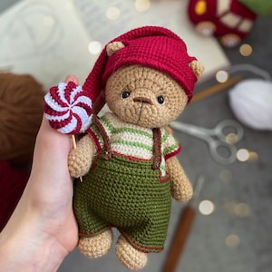 May include: A crocheted teddy bear wearing a red cap and green dungarees with brown braces. The bear holds a red and white swirled lollipop. The bear's shirt has green and white stripes. The bear is a handmade toy.
