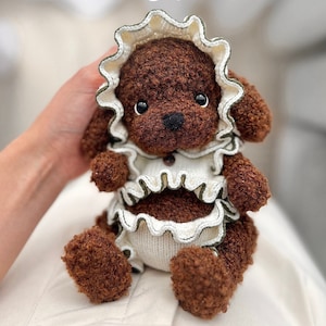 Op de afbeelding: Breipatroon voor een bruine teddybeer die een witte en groene geruite jurk draagt. De teddybeer zit op een wit oppervlak.