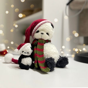 Puede incluir: Dos osos de peluche panda que llevan gorros rojos de Papá Noel y una bufanda a rayas verde y roja. El oso más grande está sentado con las piernas cruzadas. El oso más pequeño está de pie junto al más grande.