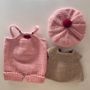 Op de afbeelding: Een roze gebreide poppenoutfit met een hartje op de voorkant, een roze gebreide baret met een rode pompon en een beige gebreide jurk.