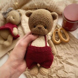May include: A brown crocheted teddy bear wearing a cream colored sweater and red overalls. The bear has black eyes and a brown nose.