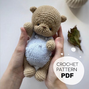 May include: A crocheted teddy bear wearing a blue knitted onesie. The bear is being held by a person's hands. The image includes the text "CROCHET PATTERN PDF".