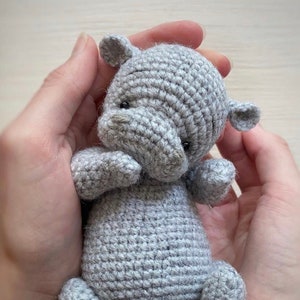 May include: A small grey crocheted stuffed animal resembling a hippopotamus. The animal has a rounded body, short legs, and a small tail.