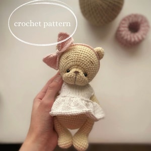 May include: A crocheted teddy bear wearing a white dress and a pink bow. The bear is light brown and has black eyes.