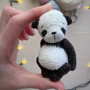 Puede incluir: Un pequeño oso panda de crochet blanco con orejas, ojos, nariz y patas negras. El panda sostiene una pequeña bola de crochet negra y blanca.