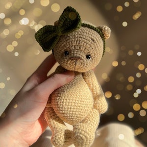 May include: A crocheted teddy bear with a green bow on its head. The bear is light brown and has black eyes. The bear is being held by a hand.