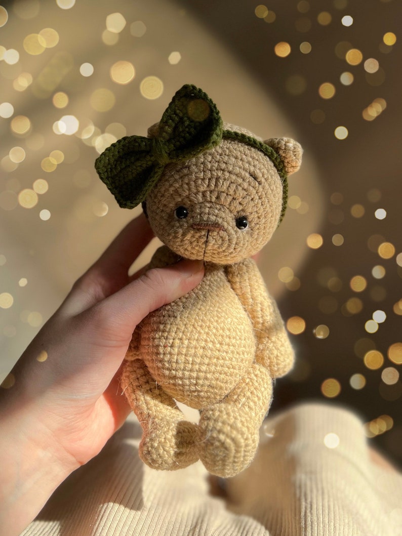 May include: A crocheted teddy bear with a green bow on its head. The bear is light brown and has black eyes. The bear is being held by a hand.