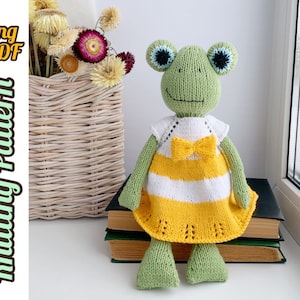 May include: Knitting pattern for a green frog wearing a yellow and white striped dress with a yellow bow. The pattern is available in English and is a PDF download.