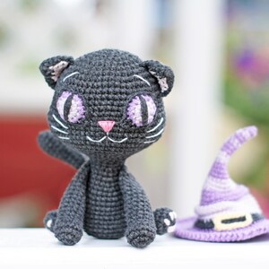 May include: A black crocheted cat with purple eyes and a pink nose. The cat is sitting on a white surface next to a purple crocheted witch hat.