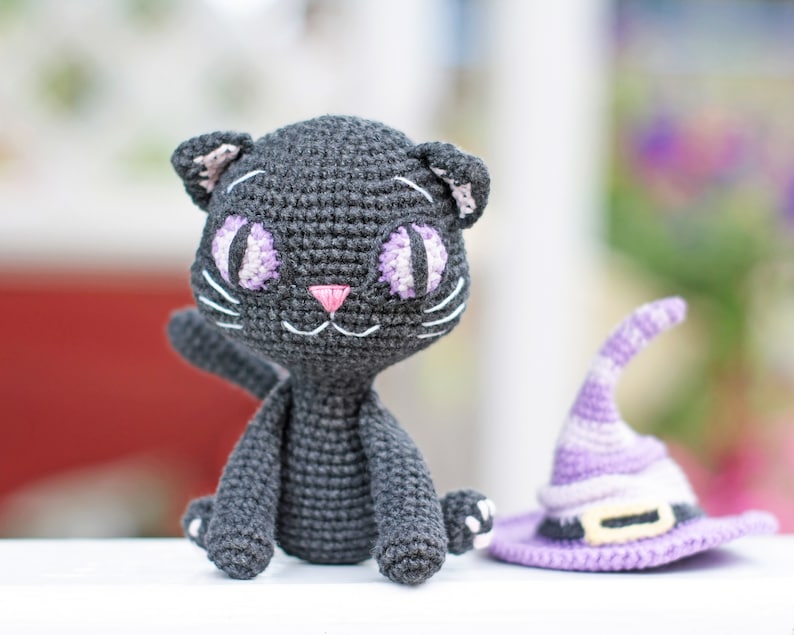 May include: A black crocheted cat with purple eyes and a pink nose. The cat is sitting on a white surface next to a purple crocheted witch hat.