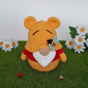 K&ouml;nnte beinhalten: Ein geh&auml;kelter Winnie Puuh-Zwerg mit einem rot-wei&szlig;en Hut und einem blauen Schmetterling auf der Nase. Der Zwerg sitzt auf einer gr&uuml;nen Grasfl&auml;che.