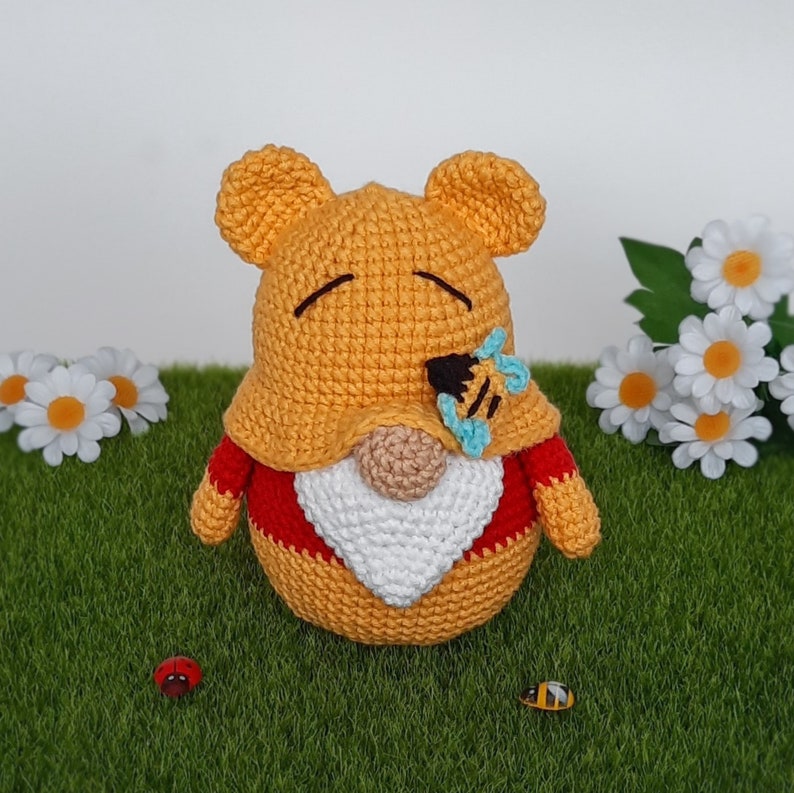 K&ouml;nnte beinhalten: Ein geh&auml;kelter Winnie Puuh-Zwerg mit einem rot-wei&szlig;en Hut und einem blauen Schmetterling auf der Nase. Der Zwerg sitzt auf einer gr&uuml;nen Grasfl&auml;che.