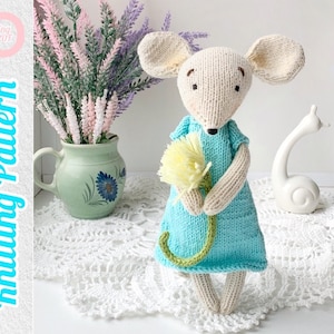 May include: Knitted mouse doll wearing a light blue dress and holding a yellow flower. The doll is sitting on a white crocheted doily.