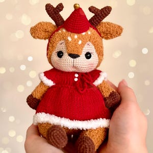 May include: A handmade crochet reindeer doll with a red dress and hat, white trim, and brown antlers. The doll has a cute, friendly expression and is held in someone's hands. The background has a bokeh effect.