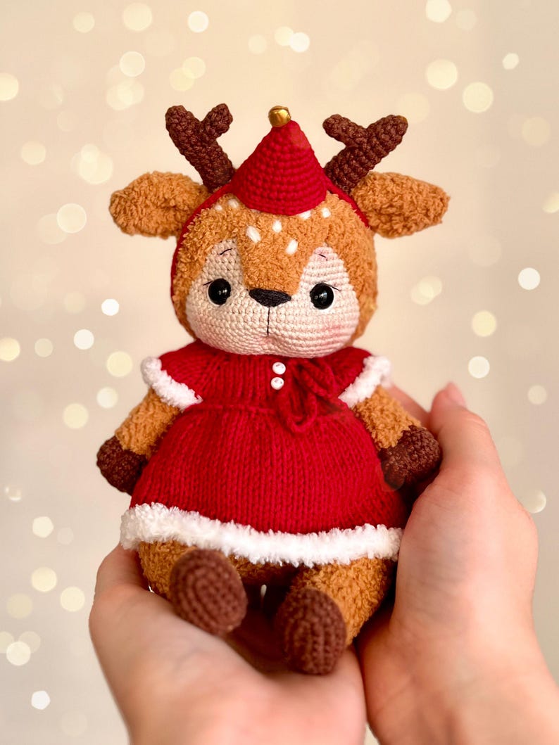 May include: A handmade crochet reindeer doll with a red dress and hat, white trim, and brown antlers. The doll has a cute, friendly expression and is held in someone's hands. The background has a bokeh effect.