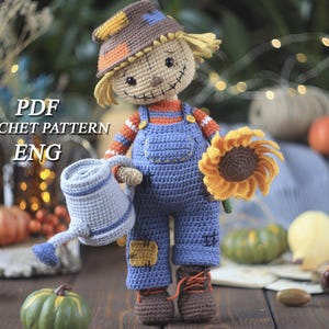 Può includere: Bambola di spaventapasseri all'uncinetto con una tuta blu, che tiene un annaffiatoio e un girasole. La bambola ha un sorriso cucito e un cappello di paglia. Il testo "PDF CROCHET PATTERN ENG" è visibile.