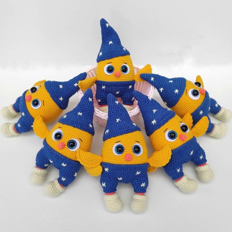 May include: Six crocheted plush toys in the shape of a yellow bird wearing blue pajamas with white stars. The birds are all wearing blue pointed hats with white stars.