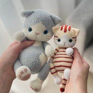 Puede incluir: Dos juguetes de crochet con forma de gato. Uno es gris con detalles blancos y el otro es blanco con rayas naranjas.