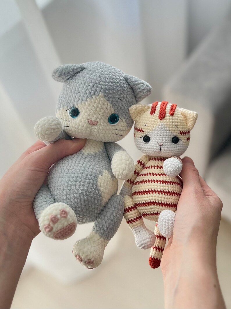 Puede incluir: Dos juguetes de crochet con forma de gato. Uno es gris con detalles blancos y el otro es blanco con rayas naranjas.