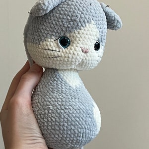 Puede incluir: Un juguete de peluche de gato gris y blanco tejido a crochet con ojos azules. El juguete est&aacute; sostenido en una mano.