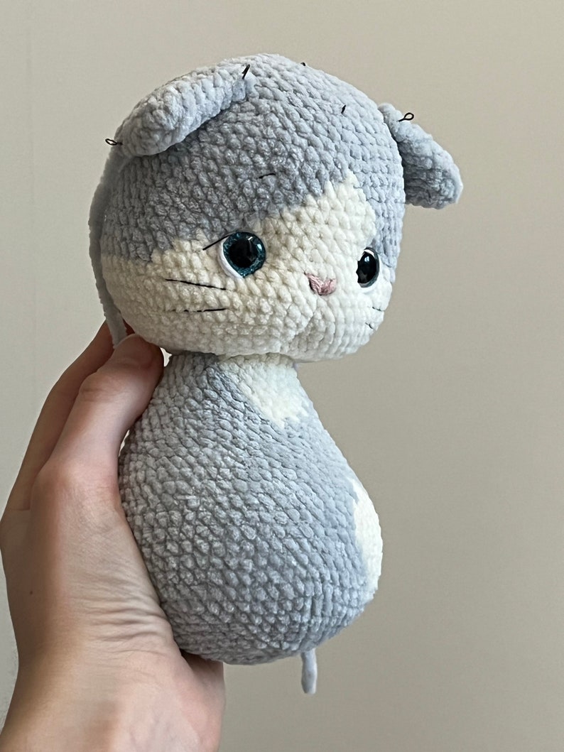 Puede incluir: Un juguete de peluche de gato gris y blanco tejido a crochet con ojos azules. El juguete est&aacute; sostenido en una mano.