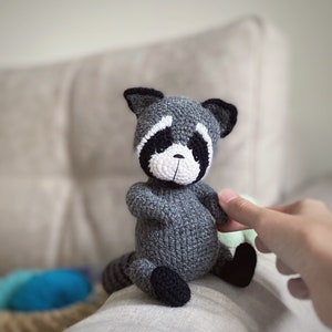 May include: A gray crocheted raccoon plush toy with black eyes, a white nose, and black ears. The toy is sitting on a white surface.