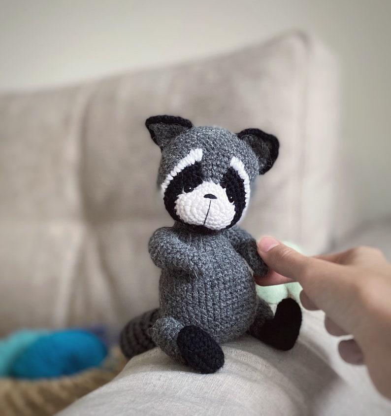May include: A gray crocheted raccoon plush toy with black eyes, a white nose, and black ears. The toy is sitting on a white surface.