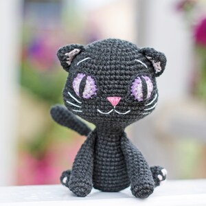 May include: A black crocheted cat toy with purple and white eyes and a pink nose. The cat is sitting on a white surface.