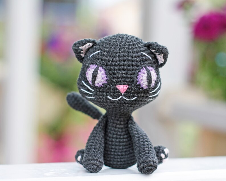 May include: A black crocheted cat toy with purple and white eyes and a pink nose. The cat is sitting on a white surface.