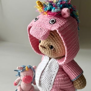 May include: A pink crocheted unicorn teddy bear wearing a pink and white unicorn costume with a rainbow mane. A smaller pink crocheted unicorn is sitting in front of the larger bear.
