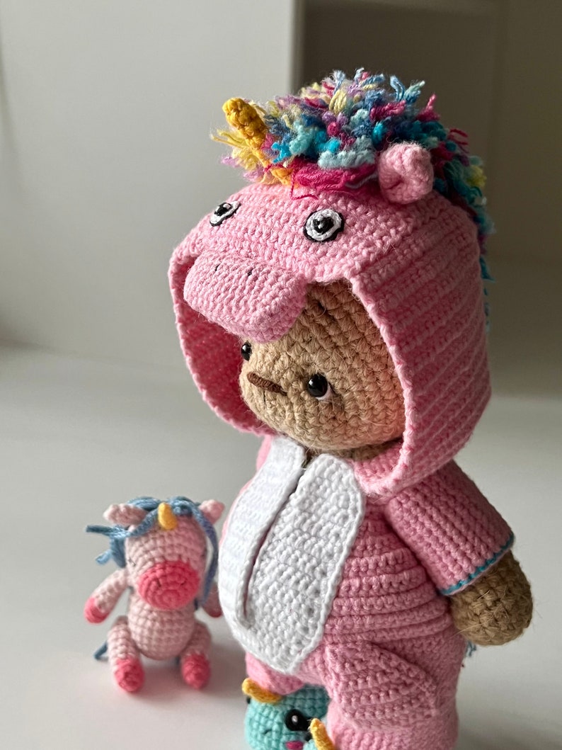 May include: A pink crocheted unicorn teddy bear wearing a pink and white unicorn costume with a rainbow mane. A smaller pink crocheted unicorn is sitting in front of the larger bear.
