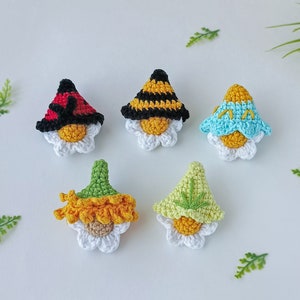 May include: Five crocheted gnome ornaments with different colored hats. The gnomes are sitting on white daisy flowers. The hats are yellow and black striped, light blue, green, and red with black spots.