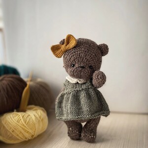 May include: A brown crocheted teddy bear wearing a green knitted dress and a yellow bow. The bear is standing on a wooden surface with yarn balls in the background.