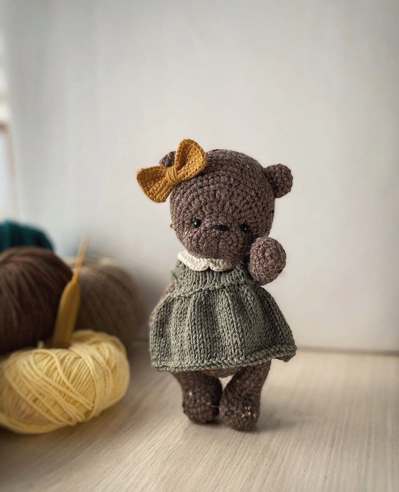 May include: A brown crocheted teddy bear wearing a green knitted dress and a yellow bow. The bear is standing on a wooden surface with yarn balls in the background.