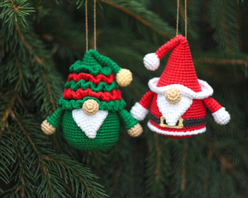 May include: Two crocheted gnome ornaments, one green with red and white accents and one red with white and black accents. Both gnomes have white beards and are hanging from a Christmas tree.