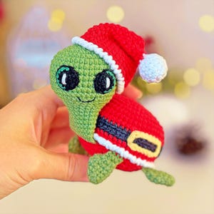 May include: A green crocheted turtle wearing a red Santa hat with a white trim and a black belt. The turtle has large black eyes and a white pom-pom on the hat.