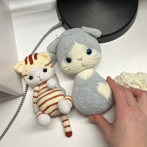 Puede incluir: Dos juguetes de gato de crochet. Uno es gris con marcas blancas y ojos azules. El otro es rayado con hilo marr&oacute;n y blanco y tiene ojos naranjas.