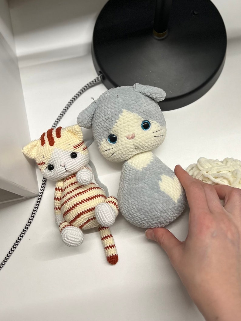 Puede incluir: Dos juguetes de gato de crochet. Uno es gris con marcas blancas y ojos azules. El otro es rayado con hilo marr&oacute;n y blanco y tiene ojos naranjas.