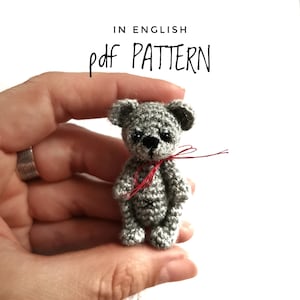 以下が含まれることがあります： 首に赤い糸が結ばれた、小さなグレーの編みぐるみテディベア。ベアは手で持たれています。ベアの上に黒い文字で「pdf PATTERN」と書かれています。