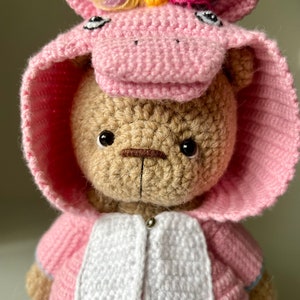 May include: A crocheted teddy bear wearing a pink unicorn costume with a rainbow mane and horn. The bear has brown eyes and a brown nose.