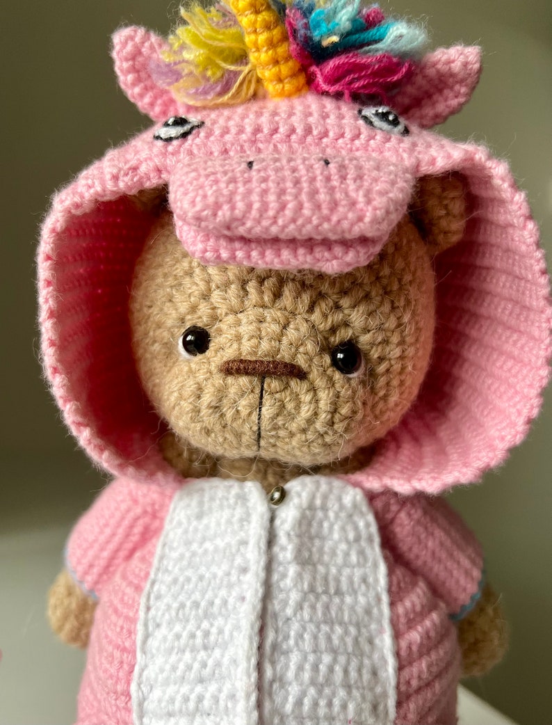 May include: A crocheted teddy bear wearing a pink unicorn costume with a rainbow mane and horn. The bear has brown eyes and a brown nose.