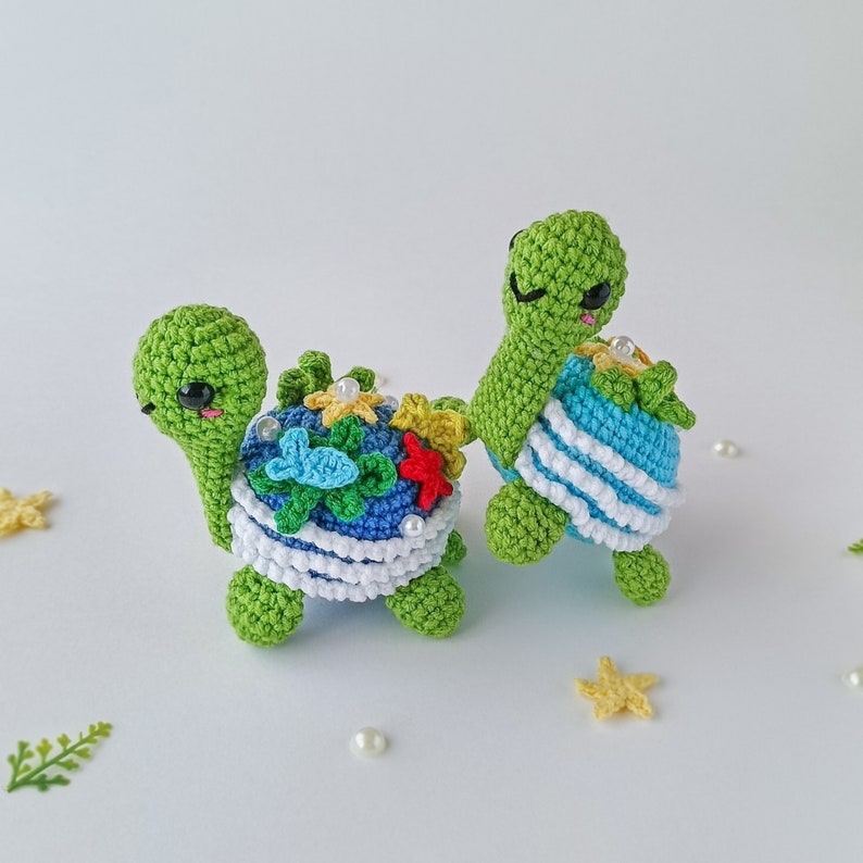 May include: Two crocheted green turtle toys with colorful details. One turtle has a blue and white shell with white stripes and the other has a blue shell with colorful flowers and a starfish.