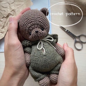 May include: A crocheted brown bear wearing a green hooded outfit. The bear is being held by a person's hands. The text "crochet pattern" is written in white on a white oval background.