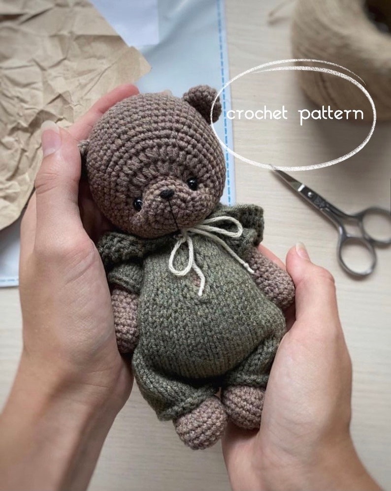 May include: A crocheted brown bear wearing a green hooded outfit. The bear is being held by a person's hands. The text "crochet pattern" is written in white on a white oval background.