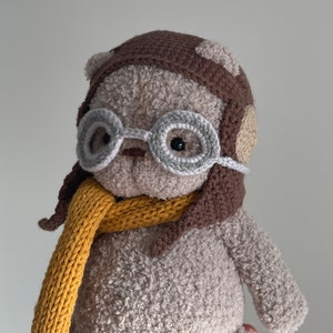 May include: A crocheted plush toy bear wearing a brown aviator hat with ear flaps, grey round glasses, and a yellow knitted scarf.