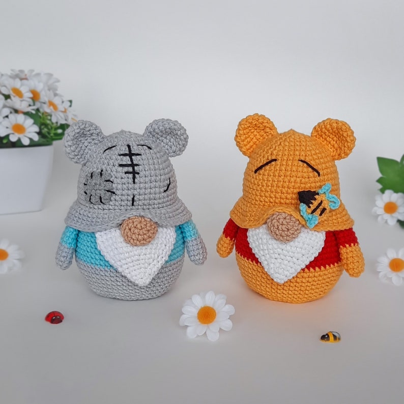 K&ouml;nnte beinhalten: Zwei geh&auml;kelte Gnom-Puppen, eine graue mit einer blau-wei&szlig;en M&uuml;tze und die andere orange mit einer rot-wei&szlig;en M&uuml;tze. Beide Gnome haben wei&szlig;e B&auml;rte und halten eine kleine geh&auml;kelte Biene.