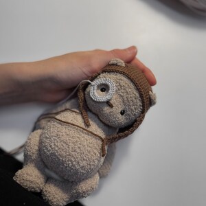 May include: A crocheted brown teddy bear with a brown and white crocheted hat. The bear has a white button eye and a black button nose.